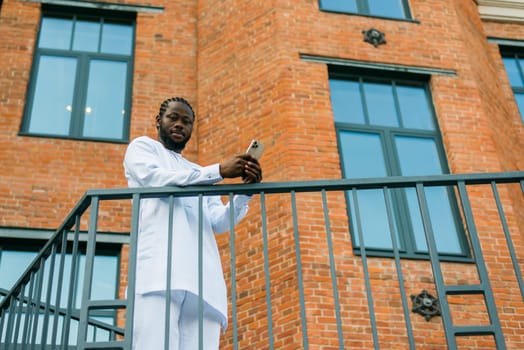 African American hipster guy in cultural and ethnic clothing dashiki chatting on smartphone outdoors, texting with friends on social networks. Millennial and Internet concept.