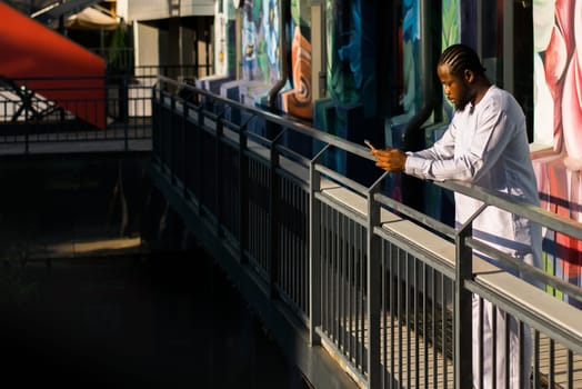 African american man in stylish ethnic dashiki wear viewing photos in social networks on mobile enjoying free time outdoors. Dark-skinned man watching funny video on smartphone and social networks. Blog and blogger scrolling content in Internet.