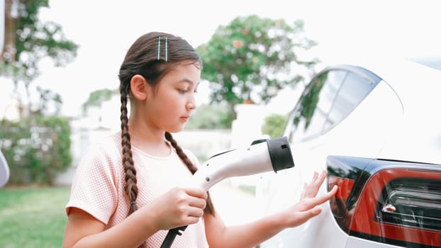 Happy little young girl learn about eco-friendly and energy sustainability as she recharge electric vehicle from home EV charging station. EV car and sustainable future generation concept. Synchronos