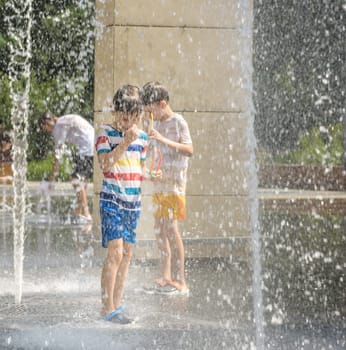 Boy having fun in water fountains. Child playing with a city fountain on hot summer day. Happy kids having fun in fountain. Summer weather. Active leisure, lifestyle and vacation.