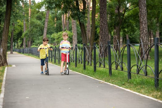 Two attractive European boys brothers, wearing red and white checkered shirts, standing on scooters in the park. They laughing, smiling, hugging and having fun. Active leisure time with kids.