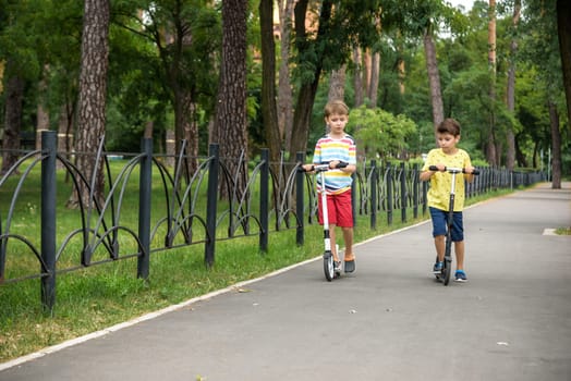 Two attractive European boys brothers, wearing red and white checkered shirts, standing on scooters in the park. They laughing, smiling, hugging and having fun. Active leisure time with kids.