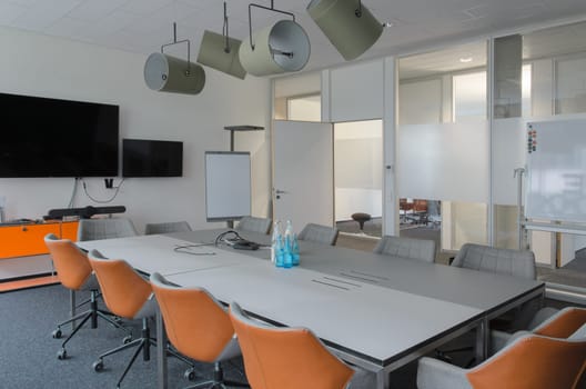 Well-lit conference room with long white table and several chairs set up for meeting. Flipchart and whiteboard and monitor tv screen are mounted on the wall at front of room. Lamps soffits on ceilling