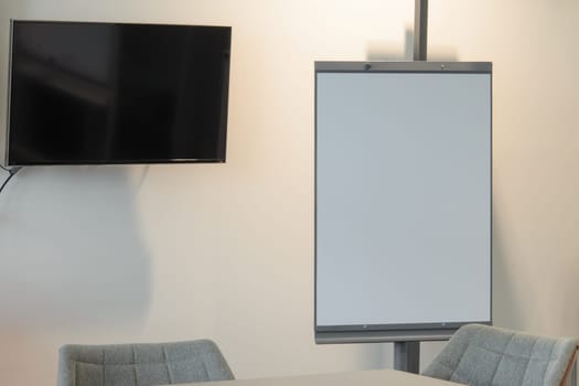 Well lit flipchart or white board in a conference room. Large black screen is cooped up on the wall. A table and chairs are partially visible in the foreground. free space, copy space