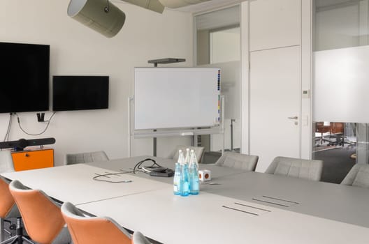 Well-lit conference room with a long white table with bottles and several chairs set up for a meeting. White board or flipchart. Monitor screens are mounted on the wall at the front of the room.