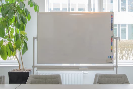 White board on window background. To the left is a green plant. Parts of a desk and chairs in the foreground.Background, place for text, copy space.
