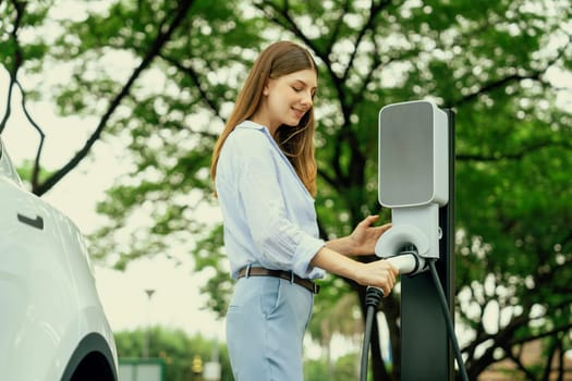 Young woman recharging battery for electric car during road trip travel EV car in natural forest or national park. Eco friendly travel during vacation and holiday. Exalt