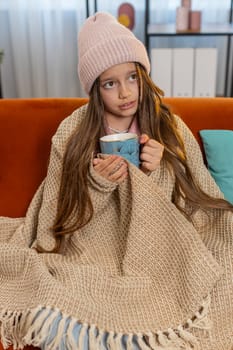 Sick ill girl child wrapped in blanket shivering from cold drinking hot herbal medical tea in unheated home apartment. Unhealthy Caucasian teenager try to warming up sitting on sofa in living room.