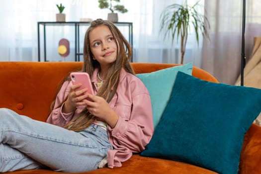 Young girl texting share messages content on smartphone social media applications online watching relax movie. Female Caucasian teenager kid child uses mobile phone sits on sofa in living room at home