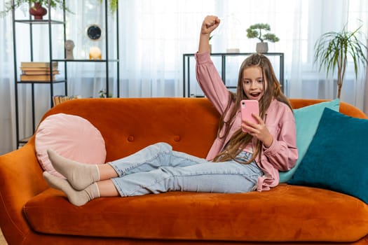 Happy girl using mobile smartphone typing playing browsing found out great big surprise, good positive news, celebrate online game win. Female teenager kid child clapping in living room sits on sofa.