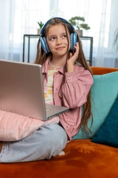 Caucasian preteen girl in headphones using laptop listens to music or lesson, distance learning. Online education. Smiling young child kid relaxing, taking a break at home apartment sitting on couch.