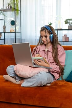 Online education at laptop of little child kid at home virtual class. Remote lesson studying of small school teen girl taking notes and viewing screen. Modern distance e-learning sits on sofa at home.