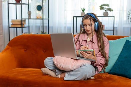 Online education at laptop of little child kid at home virtual class. Remote lesson studying of small school teen girl taking notes and viewing screen. Modern distance e-learning sits on sofa at home.