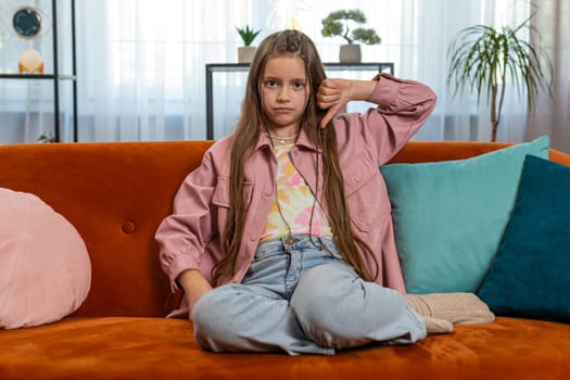 Dislike. Upset girl child showing thumbs down sign gesture, expressing discontent, disapproval, dissatisfied bad work at modern home apartment indoors. Displeased teenager kid in living room on sofa.
