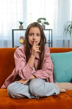Shh be quiet please. Portrait of preteen girl child presses index finger to lips makes silence hush gesture sign do not tells gossip secret. Little cute teenage female sitting on sofa in living room.