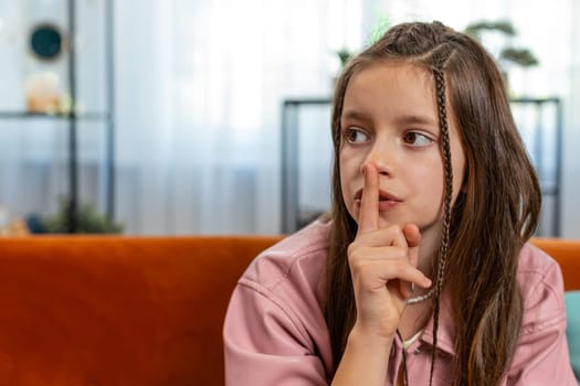 Shh be quiet please. Portrait of preteen girl child presses index finger to lips makes silence hush gesture sign do not tells gossip secret. Little cute teenage female sitting on sofa in living room.