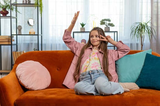 Trendy cheerful positive girl child in casual clothes having fun dancing, moving to rhythm. Teen child kid dancing emotionally rejoicing in success at home in living room sitting on sofa. Lifestyles.