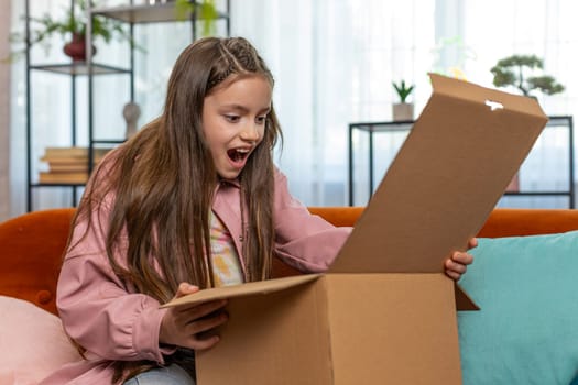 Happy amazed young child kid girl unpacking delivery parcel. Surprised satisfied teen shopper, online shop customer opening cardboard box receiving purchase gift by fast postal shipping at home.
