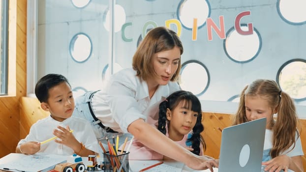 Teacher clicking at laptop while diverse student looking at screen. Children excited in presentation while drawing or writing art books at table with toys and color pencil placed on table. Erudition.