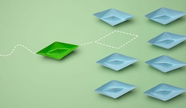 Group of blue paper boats heading in one direction and one green one heading in the opposite direction. The concept of individuality, uniqueness and talent of the employee. Get away from the influence