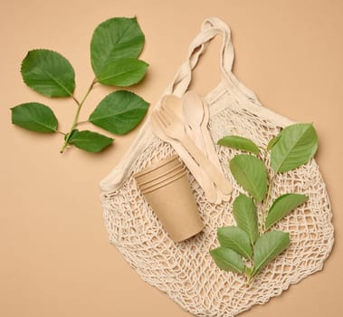 Textile knitted string bag and a stack of disposable cardboard glasses made of brown paper on a beige background and green leaves, top view