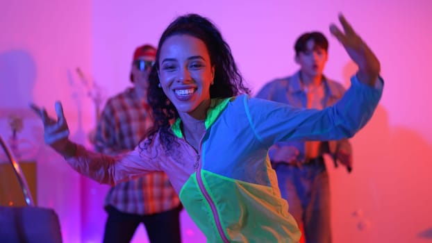 Attractive happy dancer looking at camera while smart woman moving at rhyme with neon light. Professional hispanic performer break dancing while wearing colorful cloth with diverse friend. Regalement.