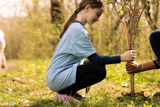 Little girl and her friend planting a tree in the ground, volunteering to the conservation project. Two teenagers taking action for reforestation, increasing vegetation in the forest, restore nature.