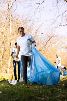African american volunteer tidying the woods area of garbage and plastic bottles, collecting trash with claw and bags. Young woman doing voluntary work to conserve natural habitat.