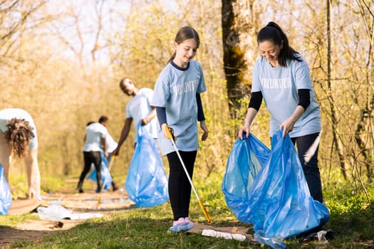 Sweet child and mother collecting trash and plastic in a disposal bag, recycling junk and cleaning the forest ecosystem. Young girl working with her mom to tidy up the nature.