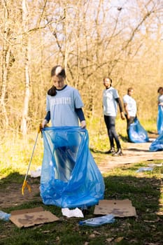 Community service volunteer reduces waste and cleans up forest setting, collecting garbage in blue trash bags. Young kid helps environment conservation by disposing of plastic waste.