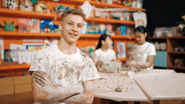 Smiling boy looking at camera and crossing arm with confident at workshop while diverse student having pottery class together. Happy caucasian student smile while pose with arm folded. Edification.