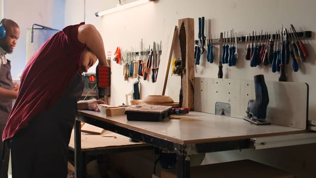 Woodworker in assembly shop using power drill to create holes for dowels in wooden board. Carpenter sinking screws into wooden surfaces with electric tool, doing precise drilling for seamless joinery, camera B