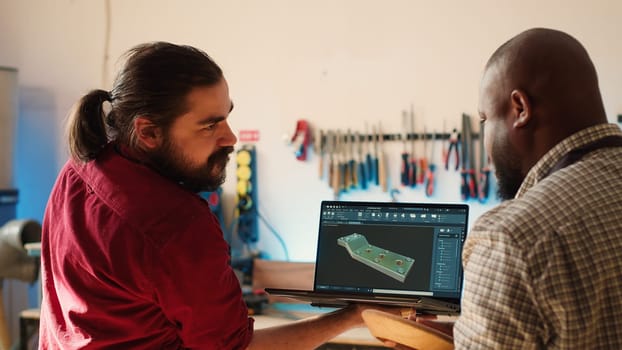 Woodworker looking over blueprints on laptop in CAD software with apprentice. Manufacturer and coworker designing custom 3D furniture on notebook in studio, preparing for customer order, camera A