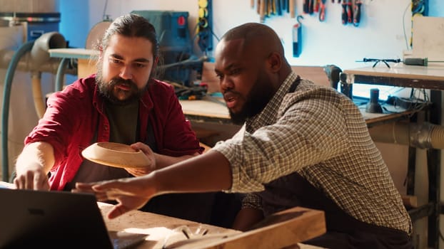 Woodworker looking over blueprints on laptop, brainstorming with african american apprentice. Team of manufacturers discussing furniture assembly schematics on notebook in studio, camera A