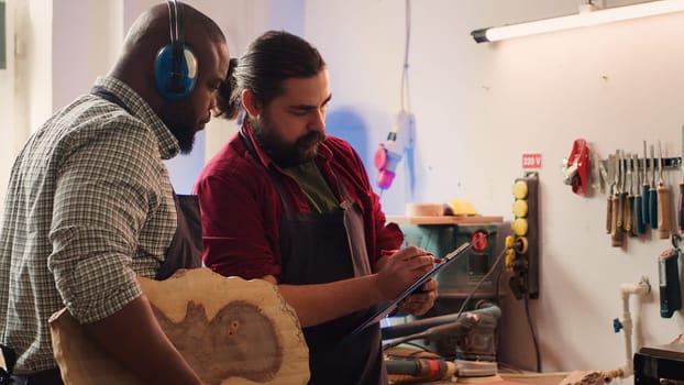 Craftsperson and colleague look over technical schematics to make commissioned wooden object. Manufacturer and coworker analyze blueprints to execute furniture assembling woodworking project, camera A