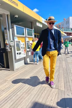 Italy, Venice, April 25, 2024, spring. A tall Italian man in yellow pants, a hat, a blue jacket and sneakers with pink laces is walking down the street. High quality photo
