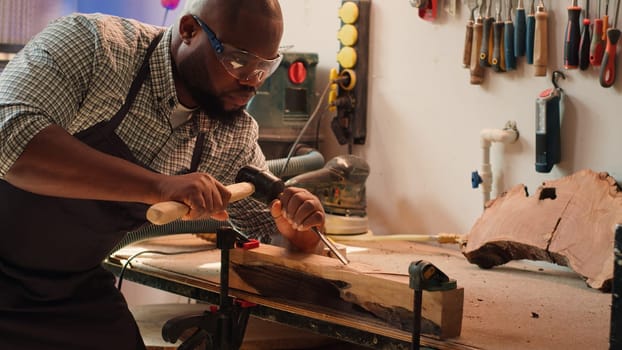 African american sculptor shaping raw timber using chisel and hammer in carpentry shop, creating wood art, wearing safety glasses. Artist making wood sculptures, engraving lumber with tools, camera A