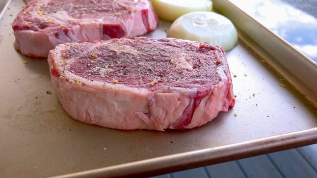 This image showcases three raw ribeye steaks, generously seasoned with coarse spices, alongside halves of fresh onions on a baking tray, prepared for a delicious grilling session.