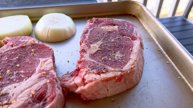 This image showcases three raw ribeye steaks, generously seasoned with coarse spices, alongside halves of fresh onions on a baking tray, prepared for a delicious grilling session.