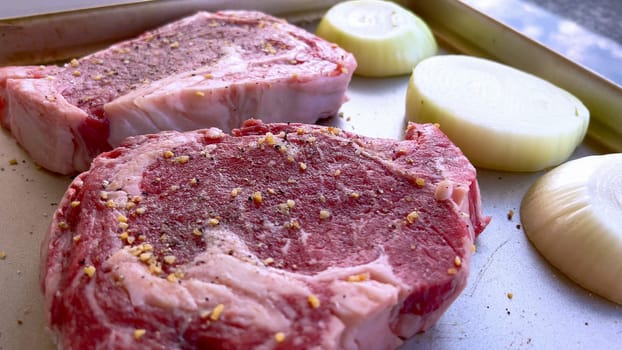 This image showcases three raw ribeye steaks, generously seasoned with coarse spices, alongside halves of fresh onions on a baking tray, prepared for a delicious grilling session.