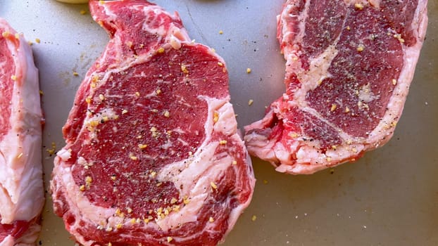 This image showcases three raw ribeye steaks, generously seasoned with coarse spices, alongside halves of fresh onions on a baking tray, prepared for a delicious grilling session.