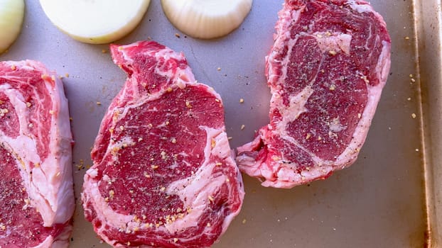 This image showcases three raw ribeye steaks, generously seasoned with coarse spices, alongside halves of fresh onions on a baking tray, prepared for a delicious grilling session.