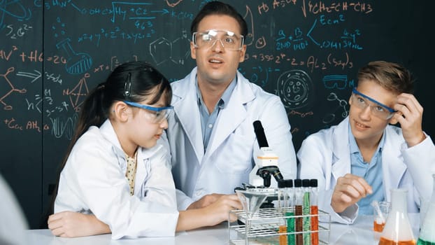 Creative teacher pointing chemistry at blackboard while talking to highschool boy at table with microscope and test tube with colored liquid. Young student wearing lab coat in STEM class. Edification.