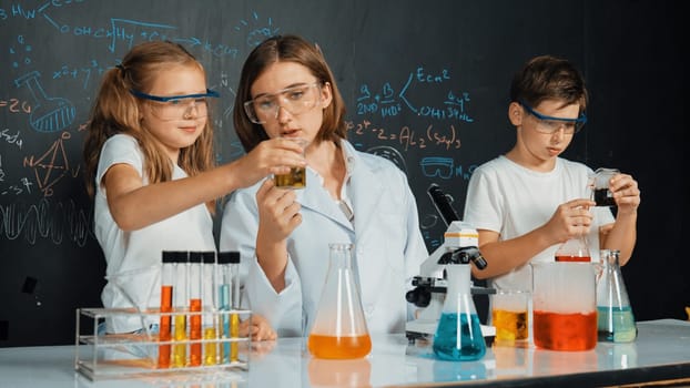 Caucasian boy mixing chemical liquid while teacher giving advice. Professional instructor wearing lab suit looking for diverse student at table with beaker filled with colored solution. Erudition.