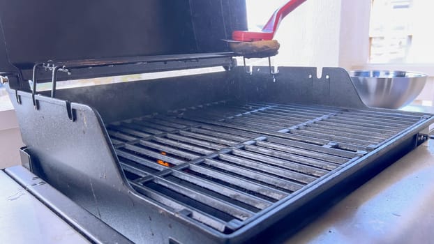 A hand uses a red grill brush to clean the black grates of a barbecue grill, ensuring it remains in perfect condition for the next grilling session.