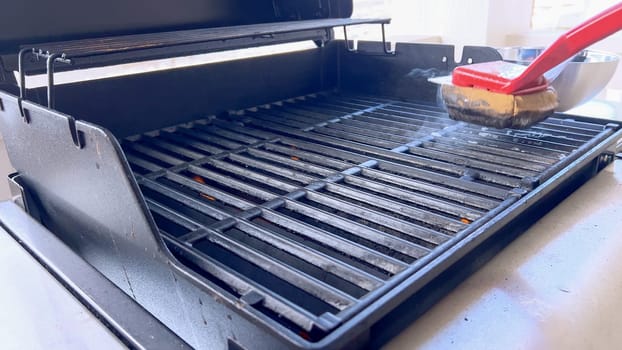 A hand uses a red grill brush to clean the black grates of a barbecue grill, ensuring it remains in perfect condition for the next grilling session.