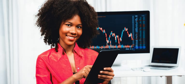 Analytical young African American businesswoman, a specialist in successful stock exchange trading, against dynamic data graph displaying marketing trend analysis on screen. Tastemaker.