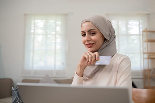 A young Muslim woman wearing a hijab sat contentedly shopping online and holding a credit card..