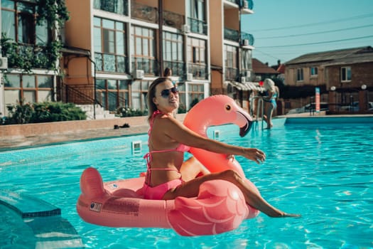 A woman is sitting on a pink flamingo float in a pool. The scene is bright and cheerful, with the woman enjoying her time in the water