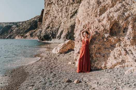 Woman red dress sea. Woman in a long red dress posing on a beach with rocks on sunny day. Girl on the nature on blue sky background